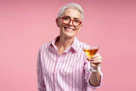 Photo for Smiling businesswoman in stylish red glasses holding cocktail shaker isolated on pink background. Attractive senior woman wearing casual pink shirt looking at camera - Royalty Free Image