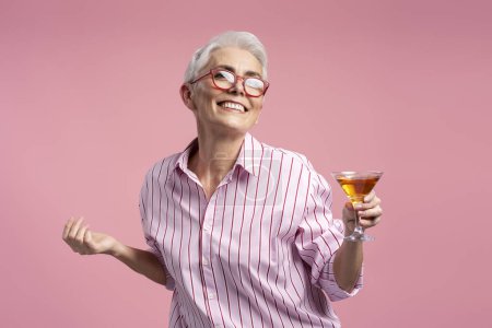 Photo for Portrait of mature gray haired female holding glass of alcohol looking at camera isolated on pink background. Smiling senior woman wearing stylish pink shirt. Concept of successful business - Royalty Free Image
