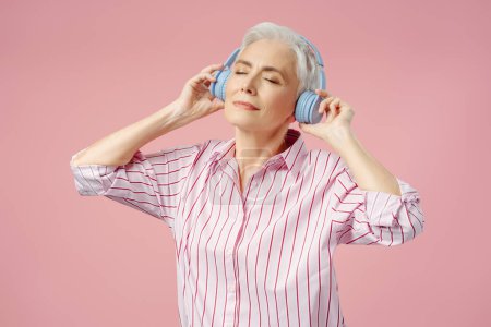 Photo for Smiling stylish senior woman with eyes closed listening music wearing wireless headphones, dancing, having fun isolated ob pink background. Technology, positive lifestyle concept - Royalty Free Image