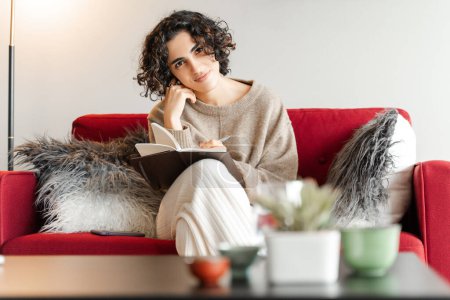 Téléchargez les photos : Belle romantique femme du Moyen-Orient écrivant des notes dans le livre laitier à la maison. Dame bouclée regardant la caméra au canapé rouge - en image libre de droit