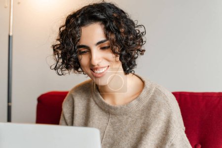 Téléchargez les photos : Femme souriante pigiste, rédactrice dactylographiant sur le clavier, utilisant un ordinateur portable fonctionnant en ligne à partir de la maison. Femme élégante portant démarrage de planification à l'appartement. Une entreprise prospère - en image libre de droit