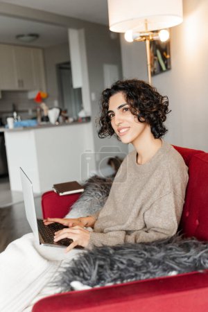 Téléchargez les photos : Belle femme souriante du Moyen-Orient en utilisant un ordinateur portable travaillant en ligne à partir de la maison. Jolie femme regardant la vidéo assis sur un canapé confortable, se détendre à la maison. Concept technologique - en image libre de droit