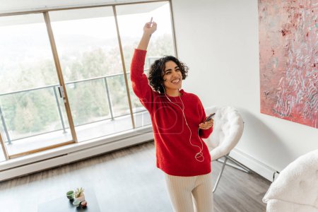Photo for Happy middle eastern woman holding mobile phone, dancing, listening favourite song at living room. Cheerful happy lady spending time at home - Royalty Free Image
