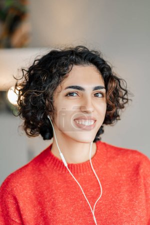 Téléchargez les photos : Portrait souriant femme du Moyen-Orient écoutant de la musique dans des écouteurs, regardant la caméra dans un appartement moderne. Technologie, concept de mode de vie positif - en image libre de droit