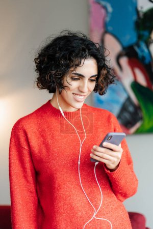 Téléchargez les photos : Vue verticale femme du Moyen-Orient souriante écoutant de la musique dans les écouteurs, regardant l'écran du smartphone dans un appartement moderne. Technologie, concept de mode de vie positif - en image libre de droit