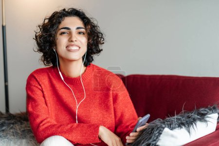 Photo for Happy young middle eastern woman wearing headphones looking at camera, enjoying favourite song, relaxing on the sofa, resting, listening calm music and daydreaming, spending lazy weekend - Royalty Free Image