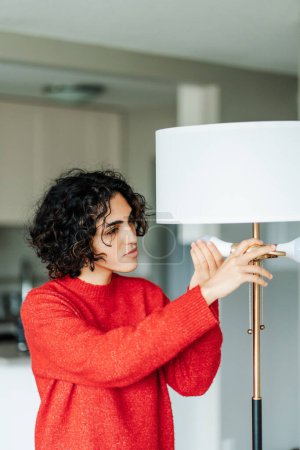 Téléchargez les photos : Curly femme du Moyen-Orient change indépendamment ampoule brûlée afin d'obtenir un éclairage de haute qualité dans la maison. Fille avec sourire insère ampoule dans le lustre - en image libre de droit