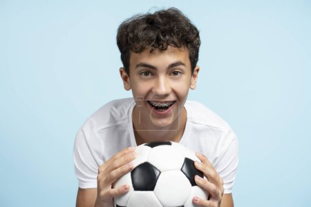 Foto de Retrato de un adolescente sonriente sosteniendo una pelota de fútbol con frenos dentales aislados sobre fondo azul, publicidad. Jugador de fútbol posando en estudio - Imagen libre de derechos