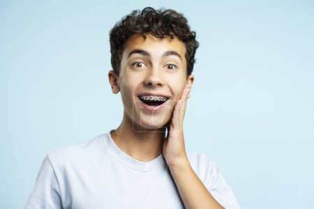 Téléchargez les photos : Adolescent excité portant des appareils dentaires touchant le visage en regardant la caméra isolée sur fond bleu. Concept de mode de vie positif - en image libre de droit
