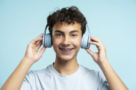 Téléchargez les photos : Portrait d'un garçon attrayant souriant avec des appareils dentaires portant des écouteurs isolés sur fond bleu. Technologie, concept publicitaire - en image libre de droit