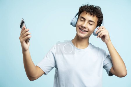 Téléchargez les photos : Portrait d'un adolescent souriant portant un casque écoutant de la musique, tenant un téléphone portable avec les yeux fermés isolés sur fond bleu. Concept technologique - en image libre de droit
