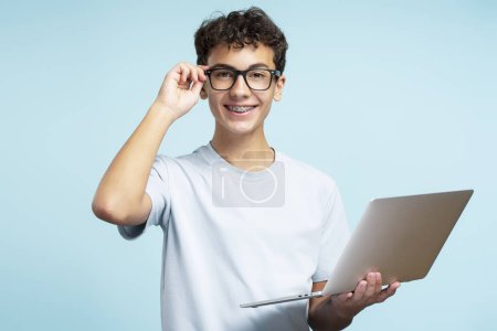 Téléchargez les photos : Portrait d'un adolescent intelligent et souriant portant des lunettes et un t-shirt décontracté regardant une caméra tenant un ordinateur portable isolé sur fond bleu. Concept technologique - en image libre de droit