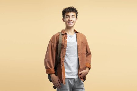 Photo for Portrait of smiling attractive teenage schoolboy with dental braces, wearing casual clothes, backpack on shoulder isolated on beige background. Education concept - Royalty Free Image