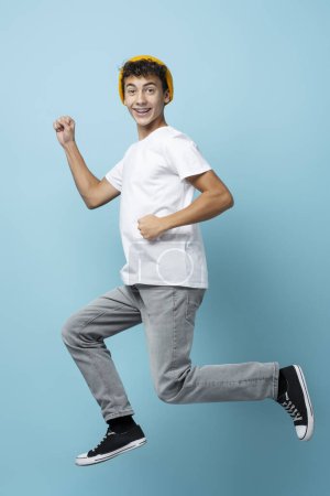 Téléchargez les photos : Portrait de garçon souriant avec chapeau jaune, sautant, isolé sur fond bleu, regardant la caméra. Concept d'enfance, hipster élégant - en image libre de droit