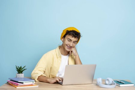 Téléchargez les photos : Adolescent attrayant souriant portant des vêtements décontractés chapeau jaune assis au bureau en utilisant le langage d'apprentissage de l'ordinateur portable isolé sur fond bleu. Concept d'éducation, de technologie - en image libre de droit