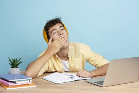 Foto de Atractivo adolescente sonriente con ropa casual bostezando sentado en el escritorio usando el ordenador portátil que se prepara para el examen aislado en el fondo azul. Concepto educativo - Imagen libre de derechos