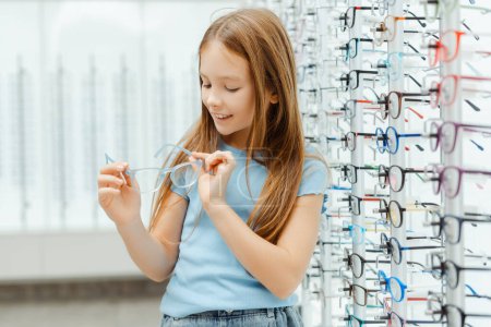 Photo for Happy little girl holding glasses while standing in the in the glasses shop. Health care concept - Royalty Free Image