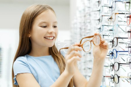 Photo for Smiling cute little girl standing in the glasses store while holding eyewear. Health care concept - Royalty Free Image