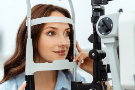 Photo for Smiling young lady examining her eyes with slit lamp in optical store. Medical examination - Royalty Free Image