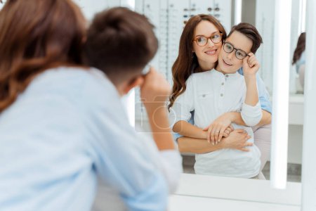 Photo for Smiling mother and her little son wearing glasses and looking at mirror in optics shop. Health care, family concept - Royalty Free Image