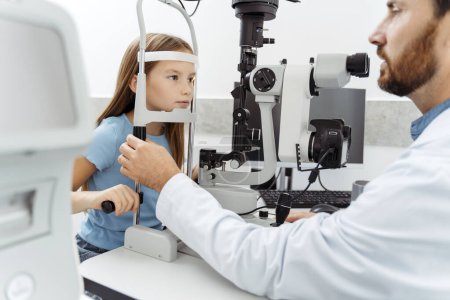 Foto de Optometrista masculino examinando a una pequeña paciente femenina con equipo oftálmico especial en óptica. Concepto de salud - Imagen libre de derechos
