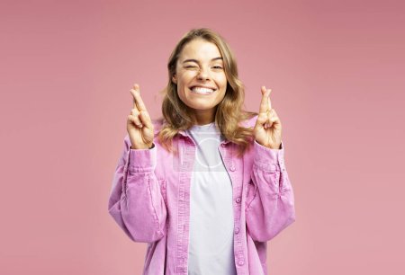 Foto de Retrato de una linda joven sonriente con una camisa casual con los dedos cruzados de pie aislados sobre un fondo rosa mirando hacia otro lado. Concepto de deseo algo, creyendo - Imagen libre de derechos