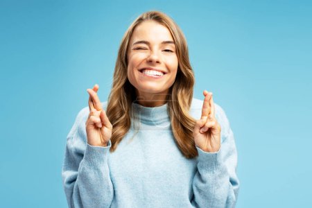 Photo for Portrait of smiling young woman wearing warm sweater with crossed fingers standing isolated on blue background looking at camera. Concept of wish something, believing - Royalty Free Image
