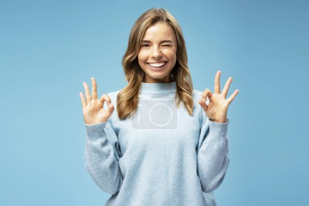 Foto de Retrato de una mujer sonriente con cabello hermoso y sedoso usando un suéter acogedor que muestra un signo aceptable mirando a la cámara de pie aislada sobre un fondo azul. Concepto de publicidad - Imagen libre de derechos
