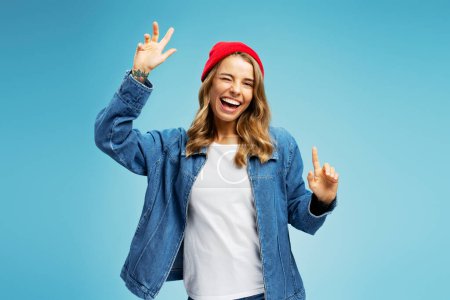 Photo for Portrait of beautiful smiling woman wearing red hat and denim shirt dancing looking at camera isolated on blue background. Concept positive fun lifestyle - Royalty Free Image