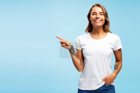 Foto de Retrato de una hermosa mujer sonriente americana con ropa casual apuntando con el dedo al espacio de copia, mirando a la cámara de pie aislada sobre un fondo azul. Concepto de publicidad, compras - Imagen libre de derechos