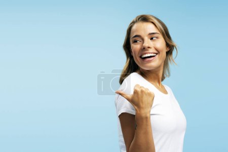Téléchargez les photos : Portrait de femme positive portant un t-shirt blanc pointant du doigt l'espace de copie, regardant loin debout isolé sur fond bleu. Concept shopping, Black Friday - en image libre de droit