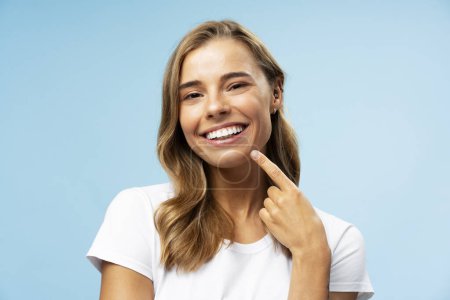 Foto de Retrato de una bella mujer sonriente señalando con el dedo sus dientes mirando a la cámara de pie aislada sobre fondo azul. Concepto de cuidado dental - Imagen libre de derechos