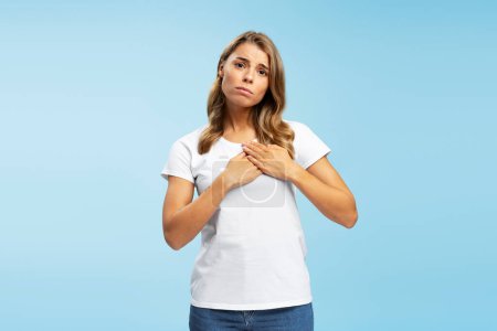 Téléchargez les photos : Portrait d'une jeune femme triste portant un t-shirt blanc décontracté tenant la main sur le cœur, faisant un geste, demandant quelque chose en regardant la caméra isolée sur fond bleu. Concept de publicité - en image libre de droit
