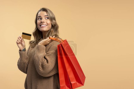 Foto de Hermosa mujer sonriente sosteniendo tarjeta de crédito y bolsas de compras rojas mirando hacia otro lado aislado sobre fondo beige, espacio de copia. Compras, venta, viernes negro, concepto de publicidad - Imagen libre de derechos