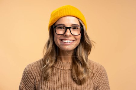 Foto de Retrato de linda hermosa mujer sonriente con gafas con estilo, sombrero hipster amarillo, suéter de invierno aislado sobre fondo beige. Estudiante universitario feliz inteligente mirando la cámara. Educación - Imagen libre de derechos
