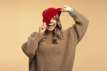 Téléchargez les photos : Portrait de belle femme heureuse met chapeau hipster rouge, portant chandail d'hiver élégant regardant la caméra isolée sur fond beige. Modèle de mode posant pour des photos en studio. Style de vie positif - en image libre de droit