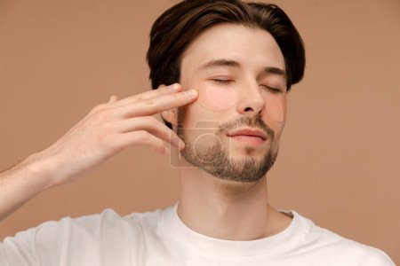 Photo for Close up of young man applying hydrogel eye patches on face. Isolated on beige background. Skin care, cosmetic, spa treatment concept - Royalty Free Image