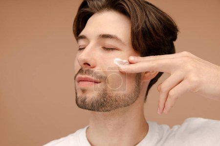 Photo for Smiling handsome young man applying moisturizer to his skin, posing in studio. Isolated on beige background. Skin care, cosmetic, spa treatment concept - Royalty Free Image