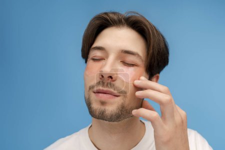 Photo for Closeup portrait of attractive metrosexual man with eyes closed applying eye patches touching face isolated on blue background. Skin care, beauty anti aging procedure, morning routine concept - Royalty Free Image