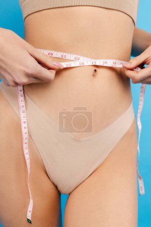 Photo for Closeup of slim young woman wearing underwear measuring her waist using measure tape isolated on blue background in studio. Fitness, diet, weight loss concept - Royalty Free Image