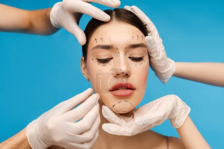 Téléchargez les photos : Cosmetologue en gants vérifiant le visage du patient avant l'opération de chirurgie esthétique. Isolé sur fond bleu. Soins de beauté, procédures anti-âge, concept de chirurgie plastique - en image libre de droit