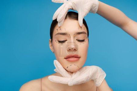 Photo for Doctor checking female face before cosmetic surgery operation. Isolated on blue background. Beauty care, anti aging procedures, plastic surgery concept - Royalty Free Image