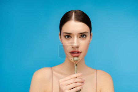 Photo for Close up of beautiful woman holding stone facial roller while massaging her chin. Isolated on blue background. Skin care, anti aging procedure - Royalty Free Image