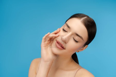 Photo for Close up of smiling young woman posing with closed eyes, touching face by her hand. Copy space. Isolated on blue background. Skin care, beauty, morning routine concept - Royalty Free Image