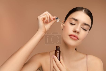 Photo for Young woman holding jar of oil serum, applying hyaluronic serum on her cheekbone. Isolated on beige background. Daily beauty treatment concept - Royalty Free Image