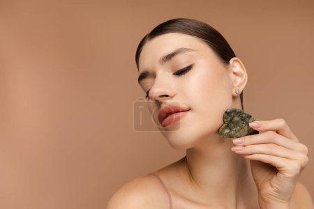 Téléchargez les photos : Belle jeune femme faisant lifting massage du visage avec gua sha grattoir en pierre. Espace de copie. Isolé sur fond beige. Soin de routine anti-âge - en image libre de droit