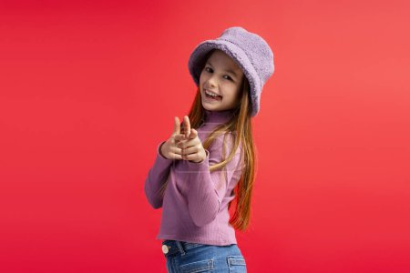 Foto de Sonriente niña alegre con elegante sombrero púrpura mirando a la cámara, señalando los dedos de pie aislados en el fondo rojo. Concepto de publicidad - Imagen libre de derechos