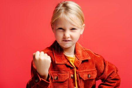 Téléchargez les photos : Portrait de petite fille en colère portant une veste marron décontractée élégante, montrant le poing, regardant la caméra isolée sur fond rouge. Concept d'émotions - en image libre de droit