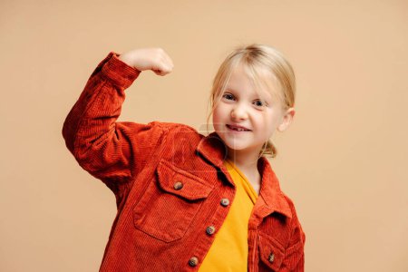 Photo for Portrait of beautiful, smiling little girl wearing casual clothes showing biceps, muscles, gesturing standing isolated on beige background. Childhood concept - Royalty Free Image