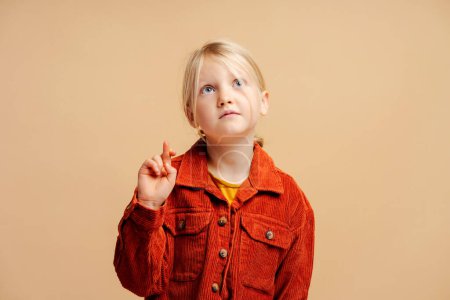 Téléchargez les photos : Portrait d'une mignonne petite fille dispendieuse portant une veste marron élégante décontractée avec les doigts croisés regardant debout isolé sur fond beige. Concept d'espoir - en image libre de droit
